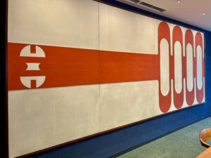 佐藤亜土作のアート作品「ユニバーサル・ポート」。白を背景に、中央を貫く太い赤の水平線と、左右に配置された幾何学的な赤い模様が特徴的な横長のモダンアートです。